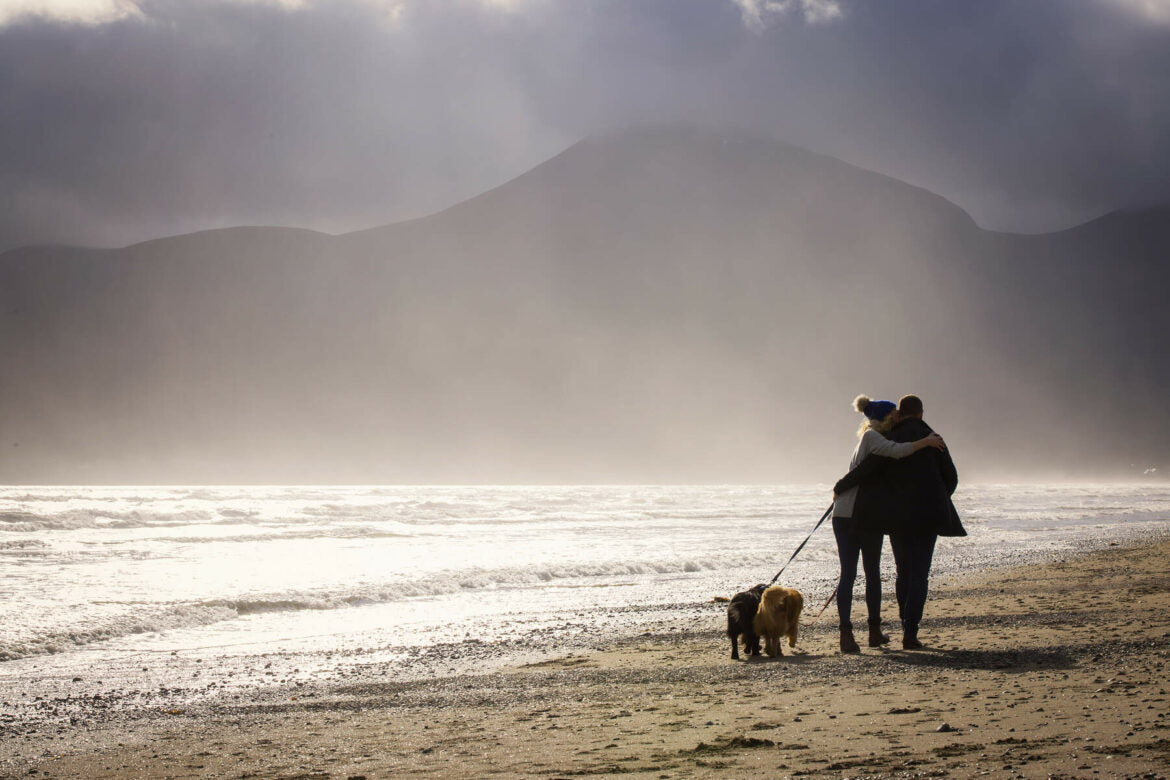 A couple stands on an Irish beach, embracing each other while walking their dog.
