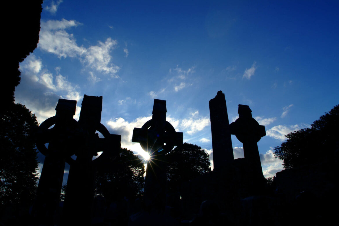 Silhouettes of several Celtic crosses against a blue sky with scattered clouds. The sun is partially hidden behind one of the crosses, illuminating the scene.