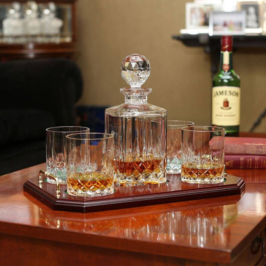 A wooden tray holds a crystal decanter and four matching glasses filled with whiskey. The setting suggests a cozy, upscale environment.
