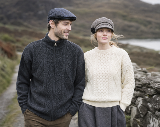 A man and woman walk along a rural path wearing knit sweaters and flat caps, with a stone wall and hills in the background.