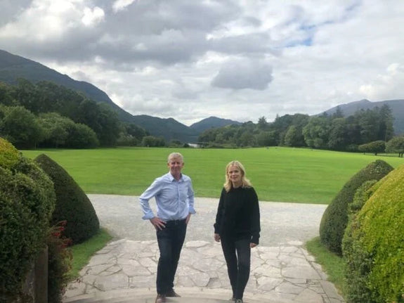John and Lulu stand on a stone path in a scenic landscape in Muckross gardens. They are facing the camera, with lush green fields and mountains in the background.
