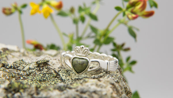 Silver Connemara Marble Claddagh Ring. A silver heart-shaped ring with a green stone sits on a textured rock surface