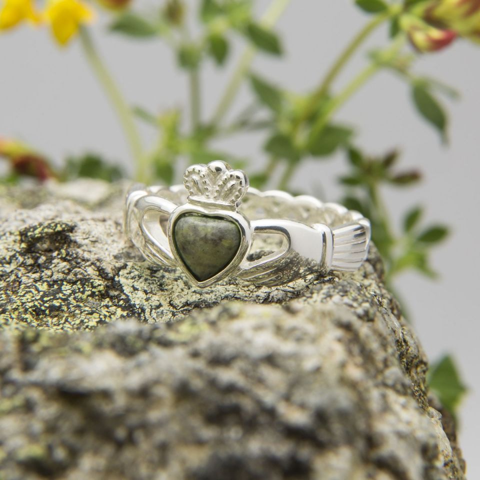 Silver Claddagh ring featuring a heart-shaped Connemara marble stone, set against a textured rock surface. Soft greenery and flowers in the blurred background add depth to the scene.