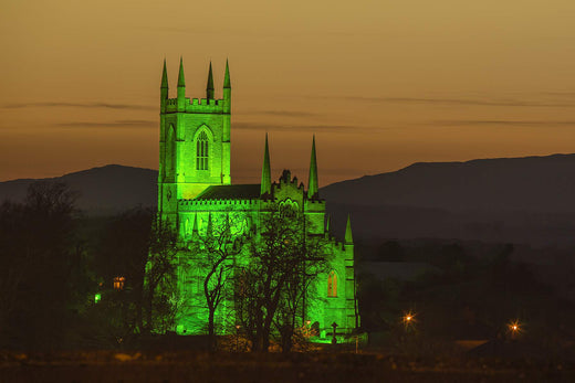 A stone church illuminated in green against a twilight sky. The building features tall spires and intricate architectural details, surrounded by dark trees and distant mountains.