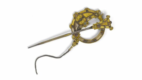 An ancient Tara Brooch with a circular base and an elongated metal arm, featuring intricate yellow designs. The brooch appears aged and weathered, suggesting historical significance.