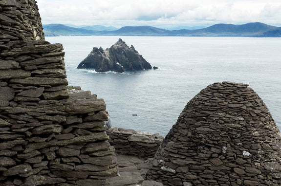 Skelligs, County Kerry. Ruins of stone structures in the foreground overlooking a calm sea. In the distance, a rocky island is visible against a backdrop of mountains and cloudy skies.