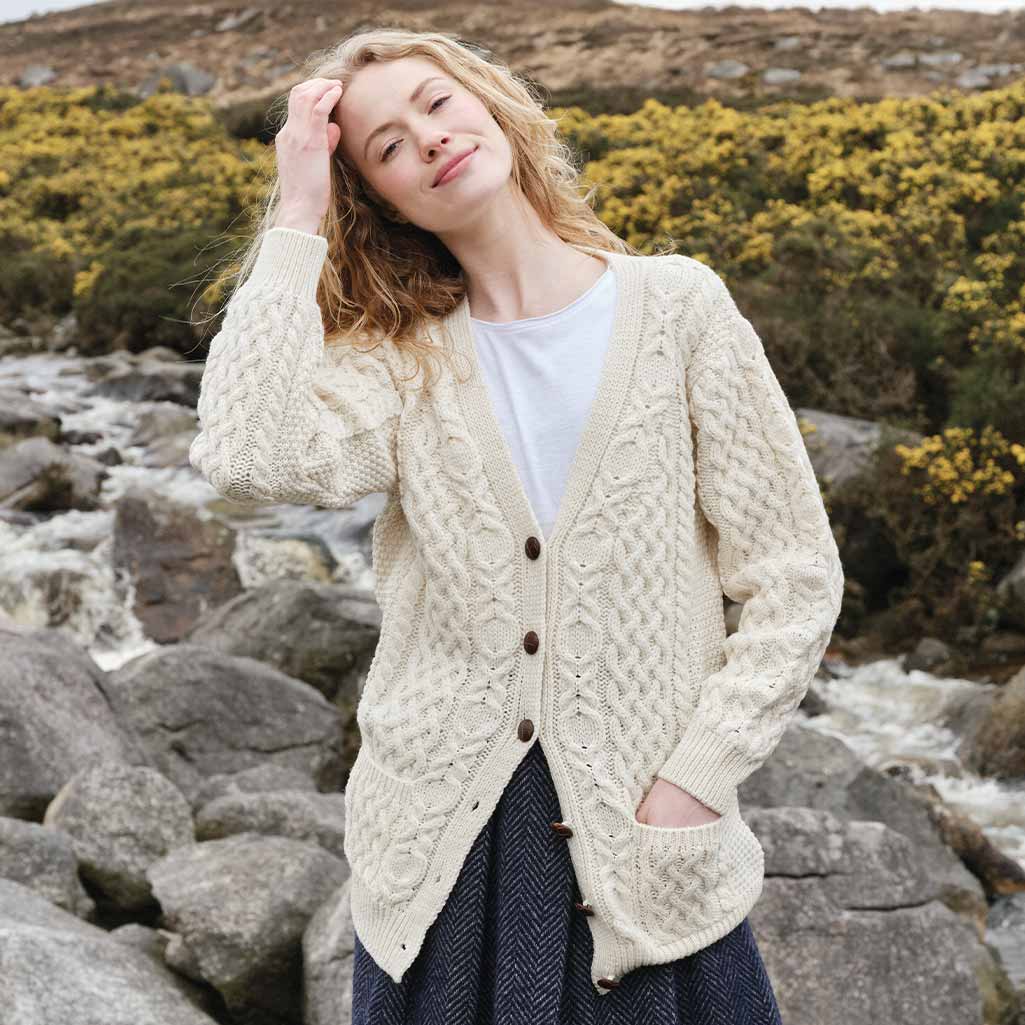 A young woman stands outdoors in a natural setting, wearing a cream-colored Aran cardigan over a white shirt. She has wavy hair and is smiling while lightly touching her hair. In the background, there are rocks and greenery, with a stream visible nearby.