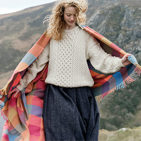 Woman wearing a traditional Aran sweater and draped in a colorful Irish wool check throw blanket from the homeware collection.
