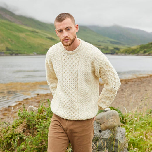 A person wearing a cream-colored cable knit sweater stands by a scenic lake and green mountains on a cloudy day.