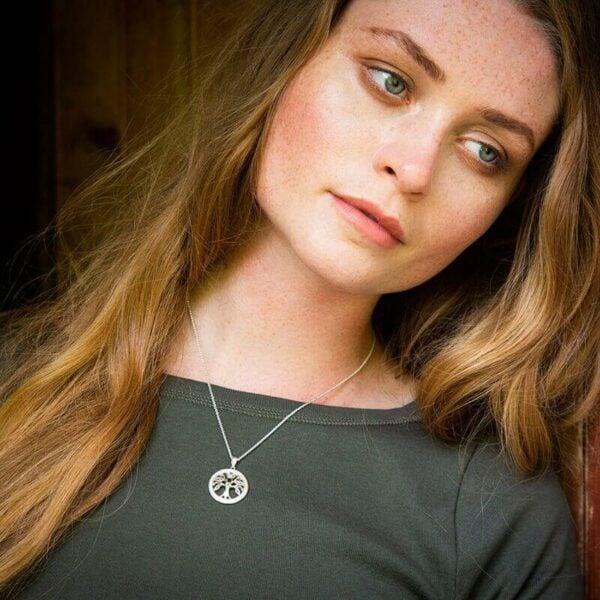 A woman with long, wavy hair gazes thoughtfully to the side. She wears a simple gray top and a silver Tree of life necklace.