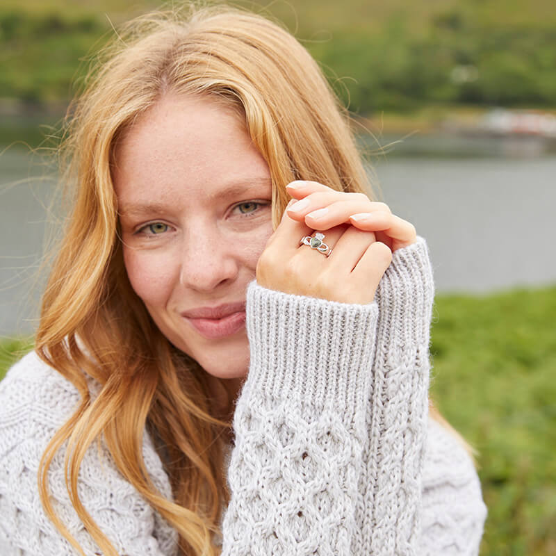 A woman with wavy blonde hair smiles softly while resting her chin on her hand. She wears a light gray knitted sweater and shows off a Claddagh Ring. The background features greenery and a body of water, suggesting an outdoor setting.