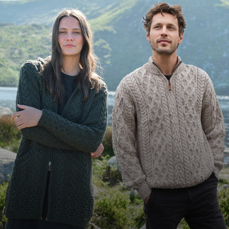 Two people modeling Aran sweaters stand outdoors near a lake with hills in the background. The woman wears a dark green sweater, while the man wears a light beige one.