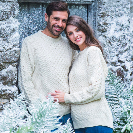 A couple stands close together, smiling in front of a rustic stone wall. They wear matching cream-colored cable-knit sweaters. Soft snow covers the ground and nearby greenery, suggesting a winter setting.