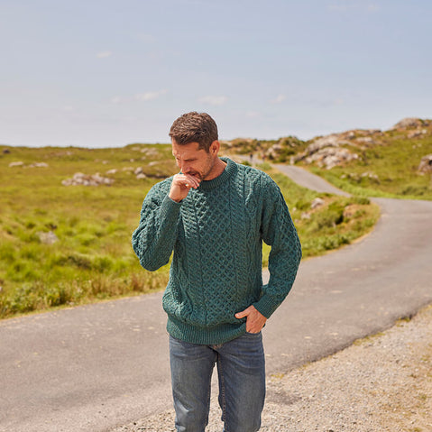 A man wearing a green crew neck Aran sweater, sitting on a stone wall with a landscape in the background.