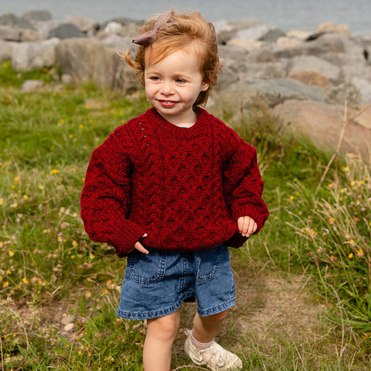 A young child walks on grass near a rocky shoreline, wearing a vibrant red kid's Aran sweater and blue denim shorts. The scene is set outdoors with greenery in the background and water visible in the distance.
