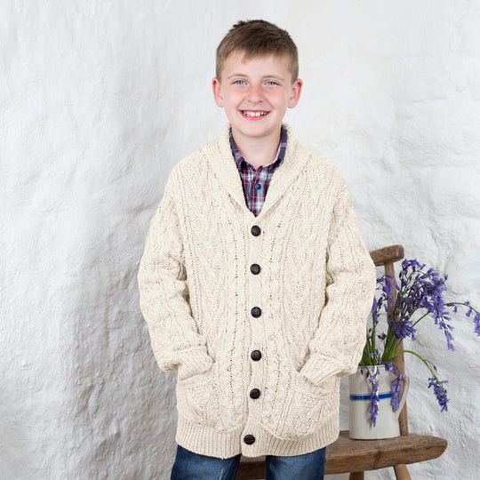 A young boy stands smiling, wearing a cream-colored kids Aran cardigan over a plaid shirt. He has his hands in the pockets of the cardigan and is positioned next to a wooden stool with a vase of purple flowers on it. The background features textured white walls.