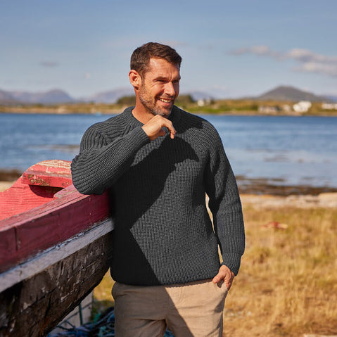 A man stands leaning on a boat by the coast wearing a traditional Irish fisherman sweater.