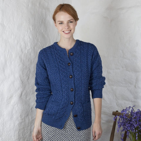 A woman standing outdoors wearing a blue cable knit Aran cardigan with button closures and a round neckline.
