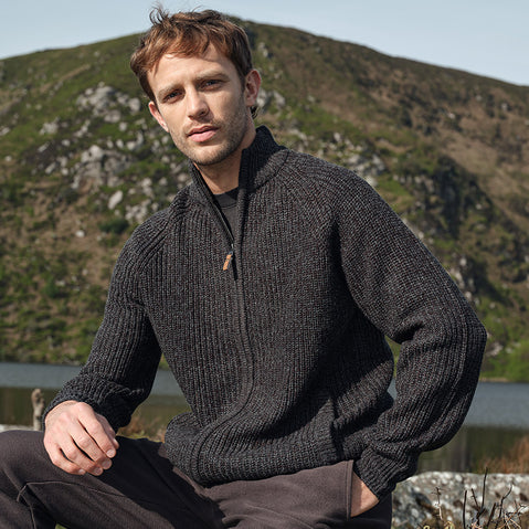 A man wearing a Men's Classic Fisherman Zipfront Wool Cardigan in Atlantic Marl sits outdoors with a scenic backdrop of hills and a lake. The dark gray wool cardigan features a zip front and chunky knit texture.