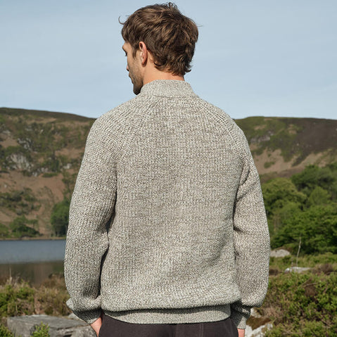 Back view of a man wearing an oatmeal-colored Men's Classic Fisherman Zipfront Wool Cardigan, standing outdoors with a scenic background of hills, greenery, and a body of water.