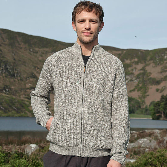 A man wearing an oatmeal-colored Irish fisherman sweater made in Ireland stands outdoors with a scenic background of hills and a lake. The cardigan is fully zipped, has a high collar, and the man's hands are in the pockets