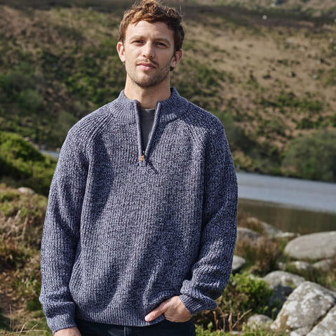 Man wearing a blue Aran sweater standing outdoors with a natural landscape in the background