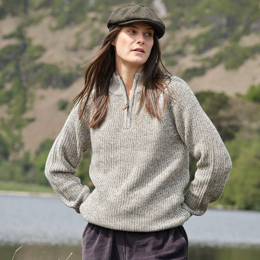 Woman wearing a knitted sweater and flat cap standing by a lake with mountains in the background