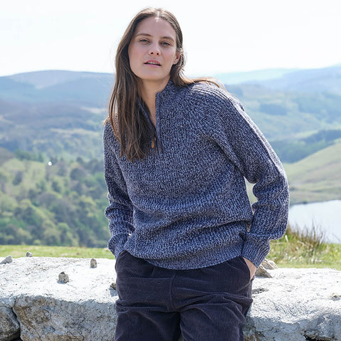 Woman wearing a blue knitted sweater and dark pants standing in a scenic outdoor setting with mountains and a lake.
