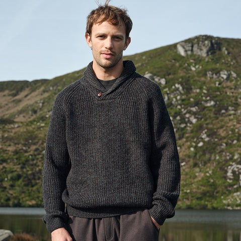 Man wearing a dark gray Aran sweater standing in front of a scenic landscape with hills and water.