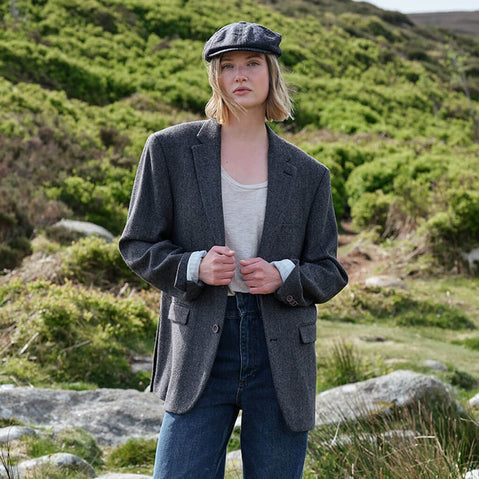 Woman wearing a gray blazer and flat cap standing in a natural landscape.