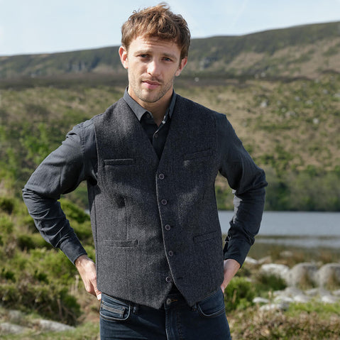Man wearing a dark gray blazer over a shirt with a vest, standing outdoors with a scenic background.