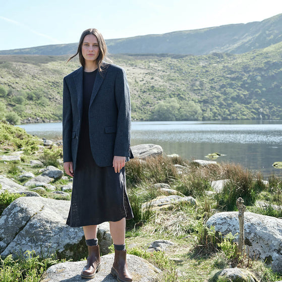 Woman in a dark coat standing on a rocky landscape with a lake and mountains in the background