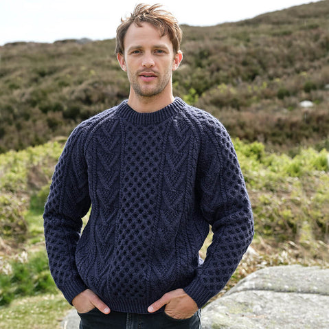 Man wearing a navy blue Aran sweater standing outdoors with a natural landscape in the background