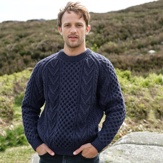 Man wearing a navy blue Aran sweater standing outdoors with a natural landscape in the background