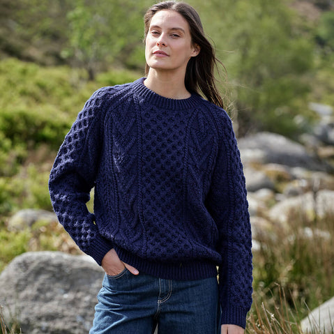 Woman wearing a dark blue Aran sweater in a natural setting with greenery and rocks.