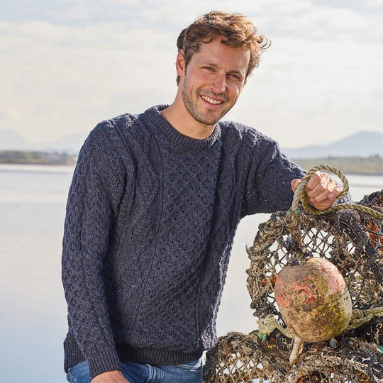 Man wearing a dark blue Aran sweater holding fishing nets by a body of water.