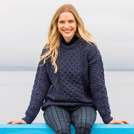 Woman wearing a dark knitted navy Aran sweater and flat cap sitting against a blurred natural background