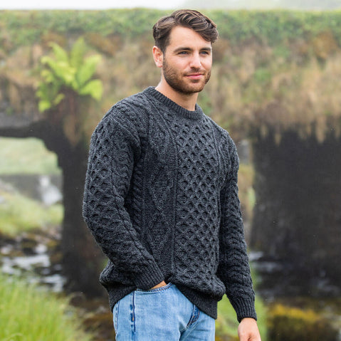 Man wearing a dark gray Aran sweater standing outdoors with greenery in the background