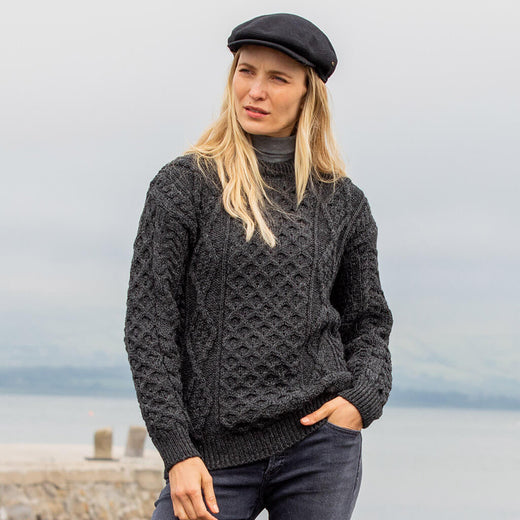 Woman wearing a dark knitted sweater and flat cap standing against a blurred natural background