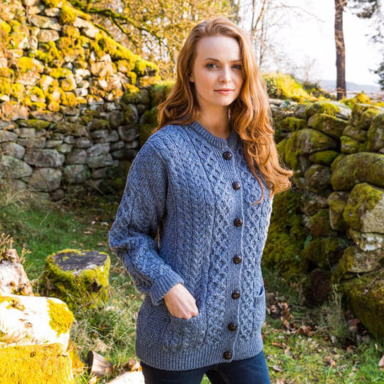 Person wearing a denim blue Women's Traditional Cable Knit Aran Cardigan, featuring intricate cable knit patterns, brown buttons, and front pockets, standing outdoors in front of a moss-covered stone wall with greenery in the background.