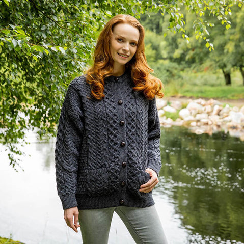 Person wearing a charcoal-colored Women's Traditional Cable Knit Aran Cardigan, featuring cable-knit patterns, front buttons, and two pockets, standing outdoors near a body of water with green foliage and trees in the background