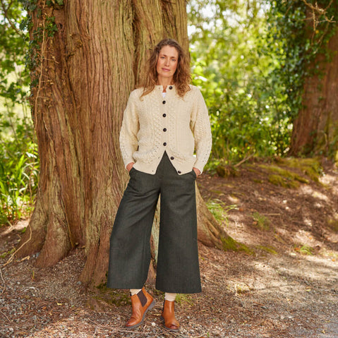 Person wearing a cream-colored Women's Traditional Cable Knit Aran Cardigan with black buttons, paired with wide-legged dark pants and brown ankle boots, standing outdoors in front of a large tree with sunlight filtering through the greenery.
