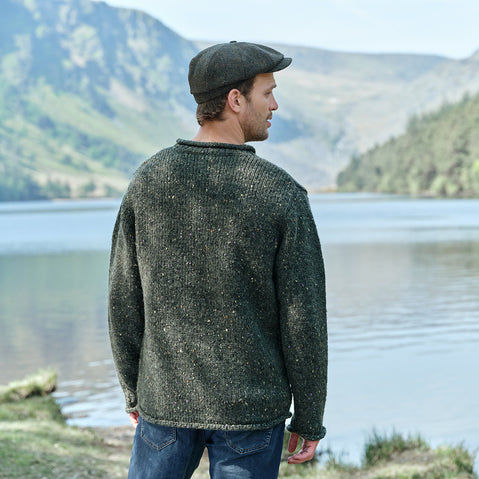 Man wearing a green sweater and cap standing by a lake with mountains in the background