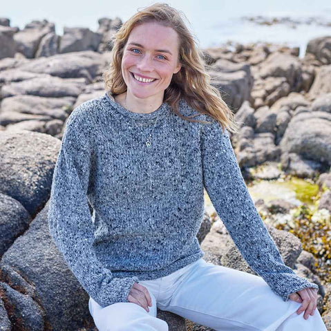 Woman wearing a grey Women's Traditional Fisherman Irish Wool Sweater sitting on rocky terrain by the sea.