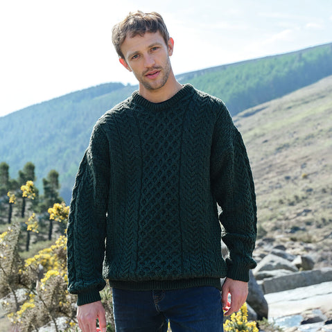 Man wearing a green sweater standing in a scenic outdoor setting with mountains and rocks.