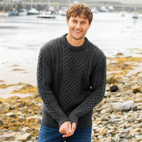Man wearing a dark charcoal gray Aran sweater standing on a rocky beach with water and boats in the background