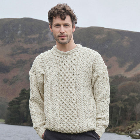 Man wearing an oatmeal colored Aran sweater standing in front of a mountainous landscape.