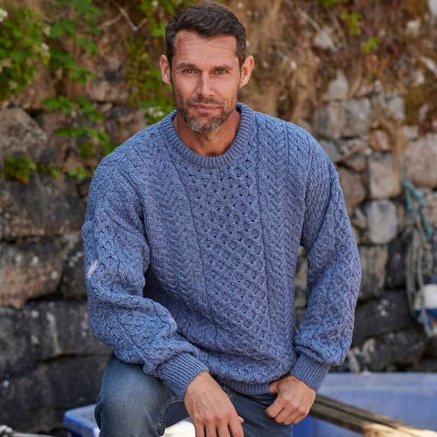 Man wearing a denim blue knitted Aransweater sitting against a stone wall.