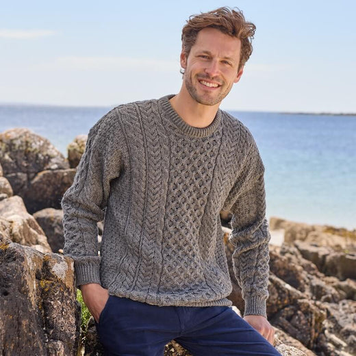 A smiling man wearing a gray cable knit Aran sweater by the ocean