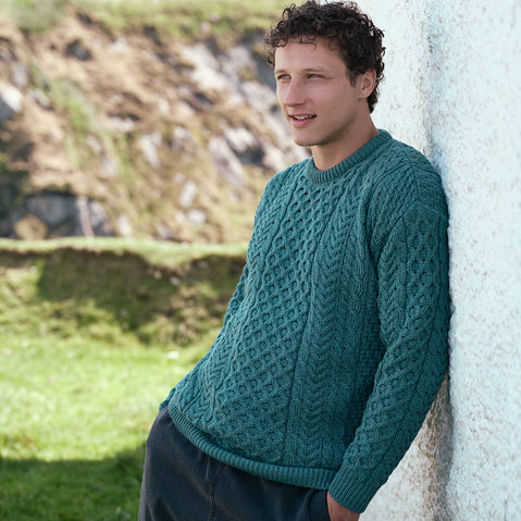 Men's Traditional Crew Neck Aran Sweater in Teal, featuring authentic Irish cable knit craftsmanship and timeless Aran stitching, styled outdoors against a rugged Irish coastal landscape.